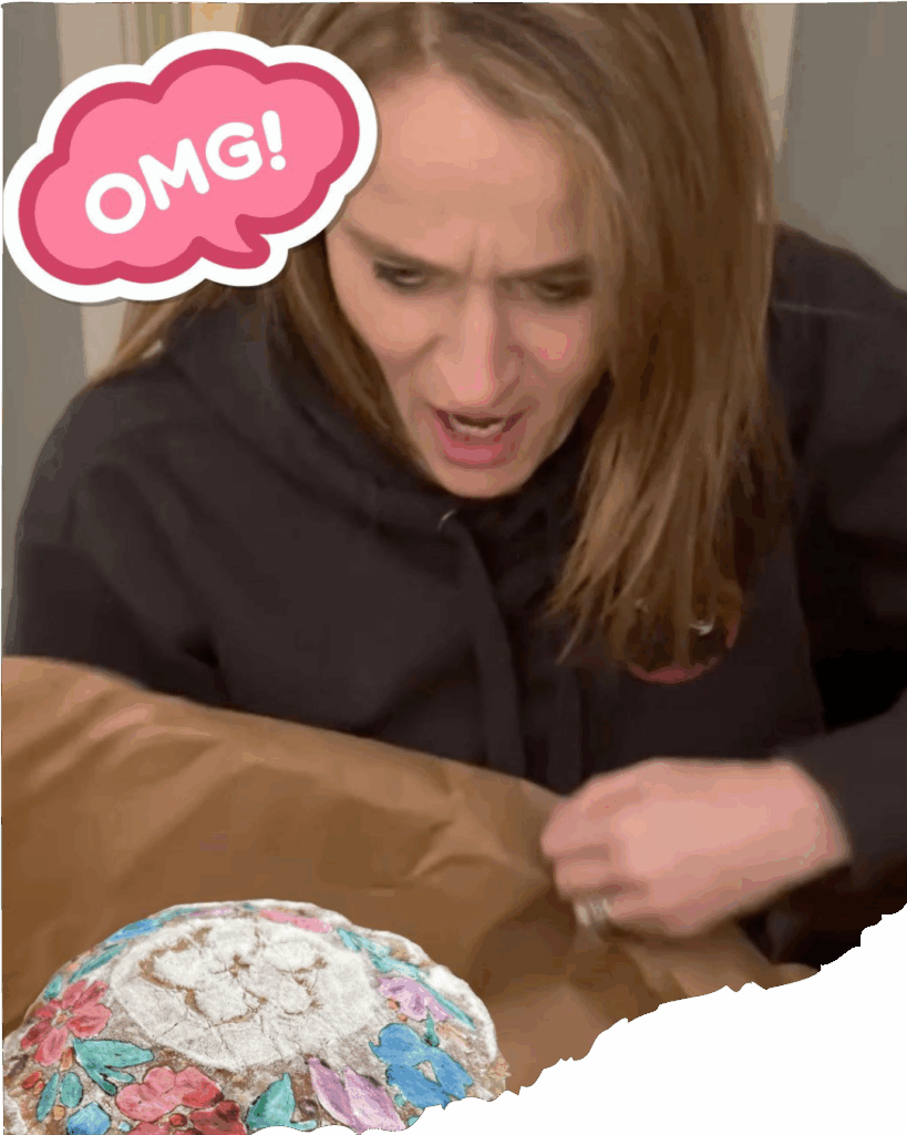 jess-omg-2 Woman gasping with surprise and delight while unboxing a painted loaf of bread decorated with colorful flowers.